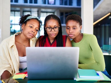 Front view of three African student girls using a laptop together, concentrated studying for a school project
